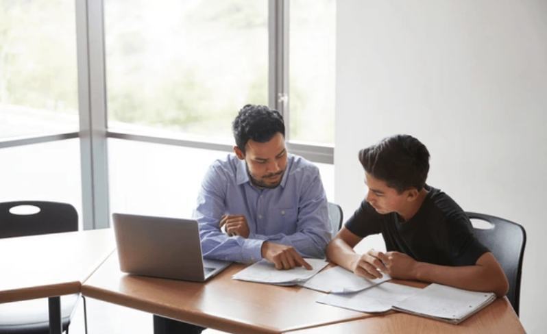 An adult and a coach seated across from each other in a bright office, working through a printed weekly planning sheet together.